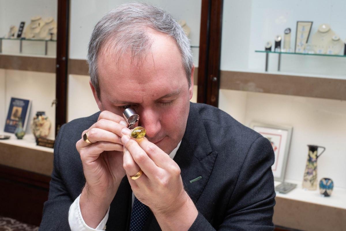 Chris Ellis at the bench in Bury St Edmunds inspecting a yellow oval gemstone ring through a jeweller’s loupe