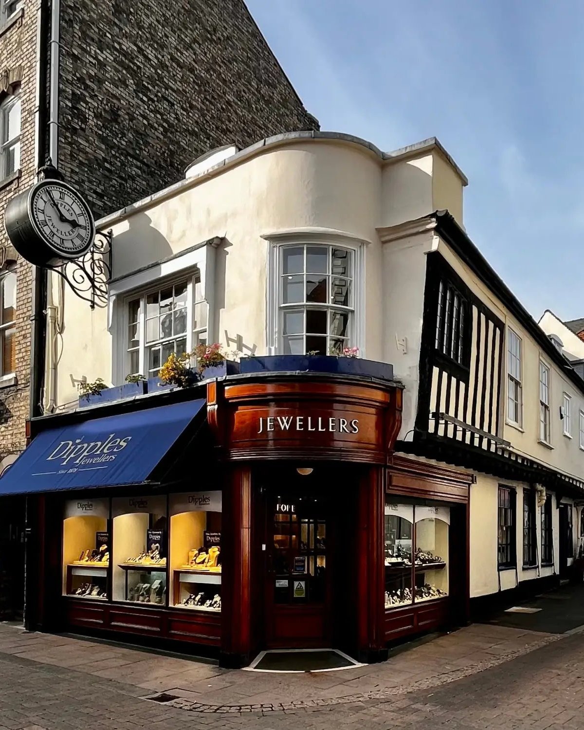 The curved walnut shopfront of Dipples Fine Jewellers at 14 Abbeygate Street, Bury St Edmunds, with the brass JEWELLERS lockup over the bow window and the projecting antique street clock on a wrought-iron bracket